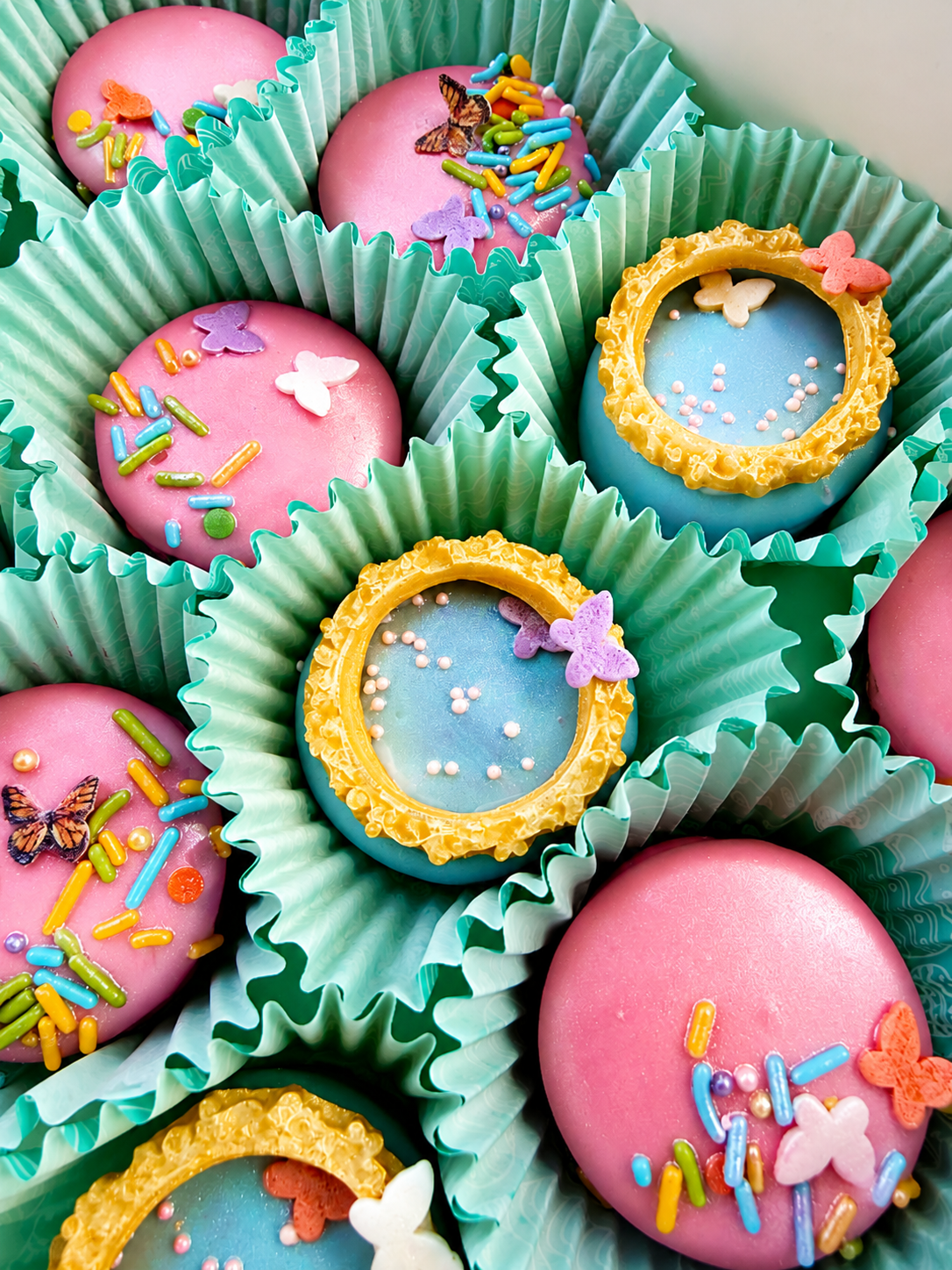 Vibrant pink and blue dipped cookies and Oreos with butterfly sprinkles and gold ornate frames in mint green liners
